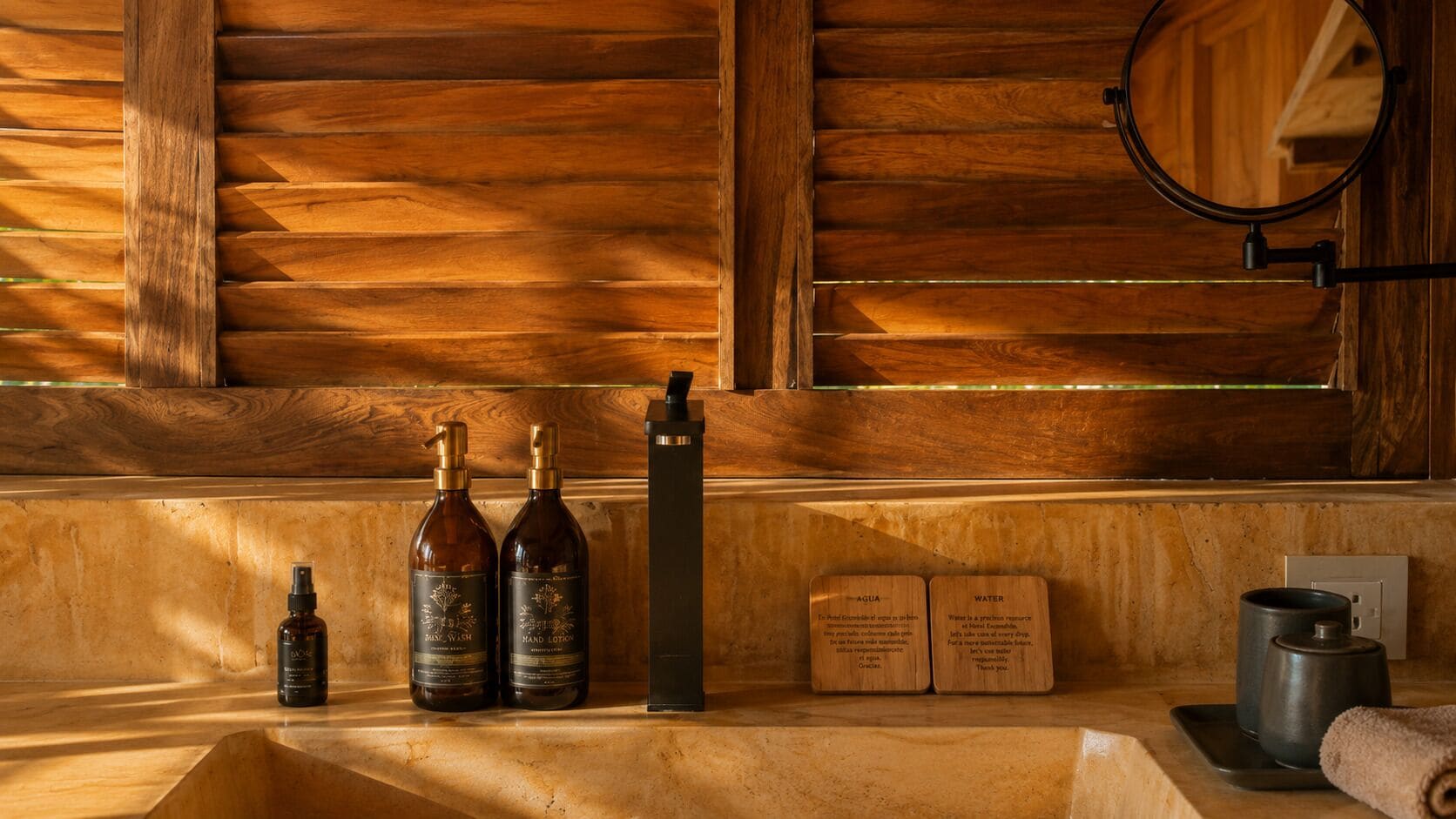 A wooden wall with a wooden shelf holding a hand lotion bottle, a hand soap bottle, a black rectangular object, and two wooden cards.