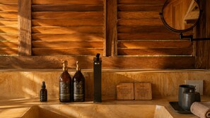 A wooden wall with a wooden shelf holding a hand lotion bottle, a hand soap bottle, a black rectangular object, and two wooden cards. - Hotel Kymaia, WorldHotels Crafted (Santa Maria Colotepec)