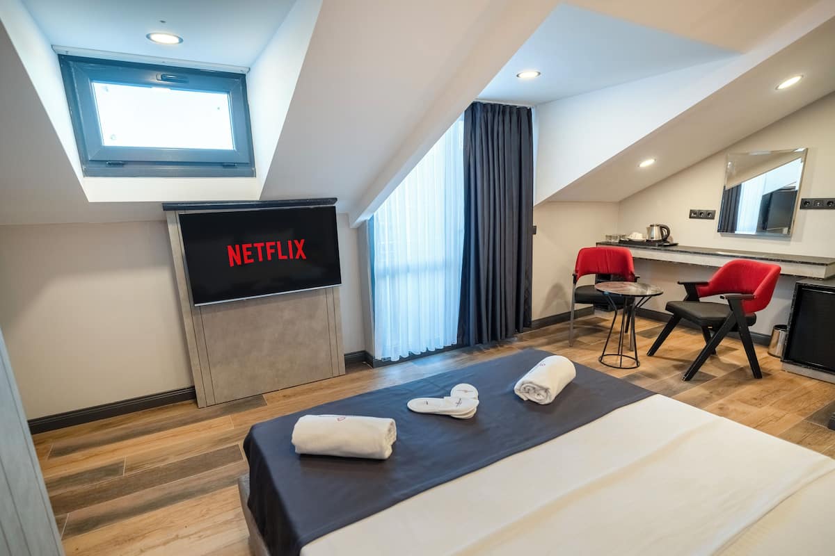 Quarto de hotel moderno com cama, escrivaninha, cadeira, TV exibindo "NETFLIX" e janela com cortinas.