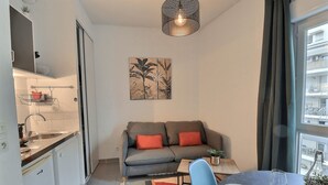 A compact living space with a kitchenette, a grey sofa with red cushions, a dining table with chairs, and a large window offering a view of the city. - Lovely apartment for 2 in Marseille (Marseille)