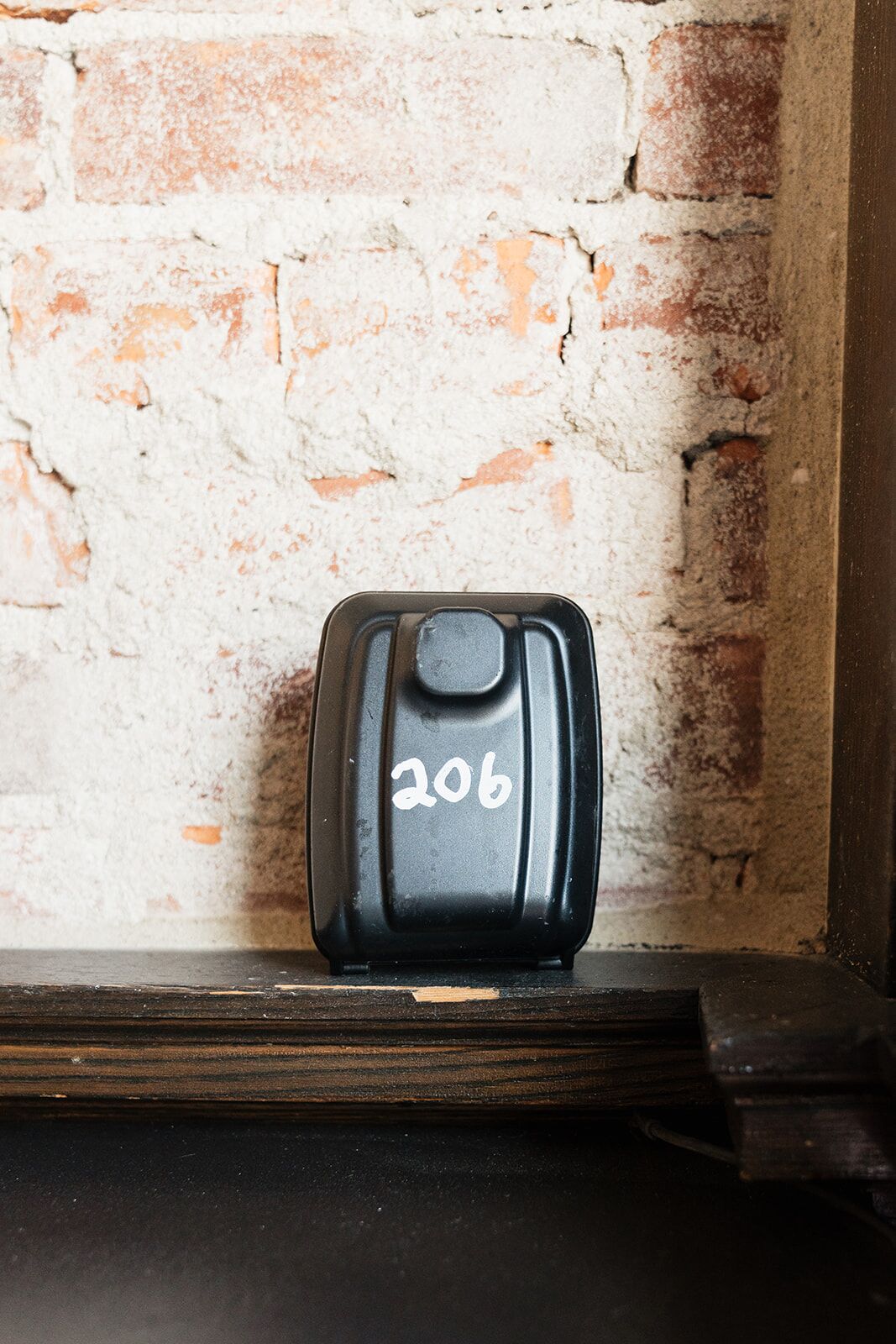 A black object with the number 206 on it, placed on a wooden ledge against a brick wall.