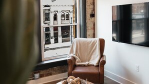 A cozy living room with a large window, a brown leather chair, a woven basket on a wooden coffee table, and a flat-screen TV mounted on the wall.