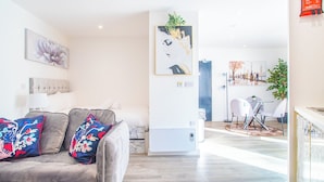 Un salon moderne avec un canapé gris, des coussins à motifs floraux, une grande peinture abstraite et une œuvre d’art représentant un paysage urbain.