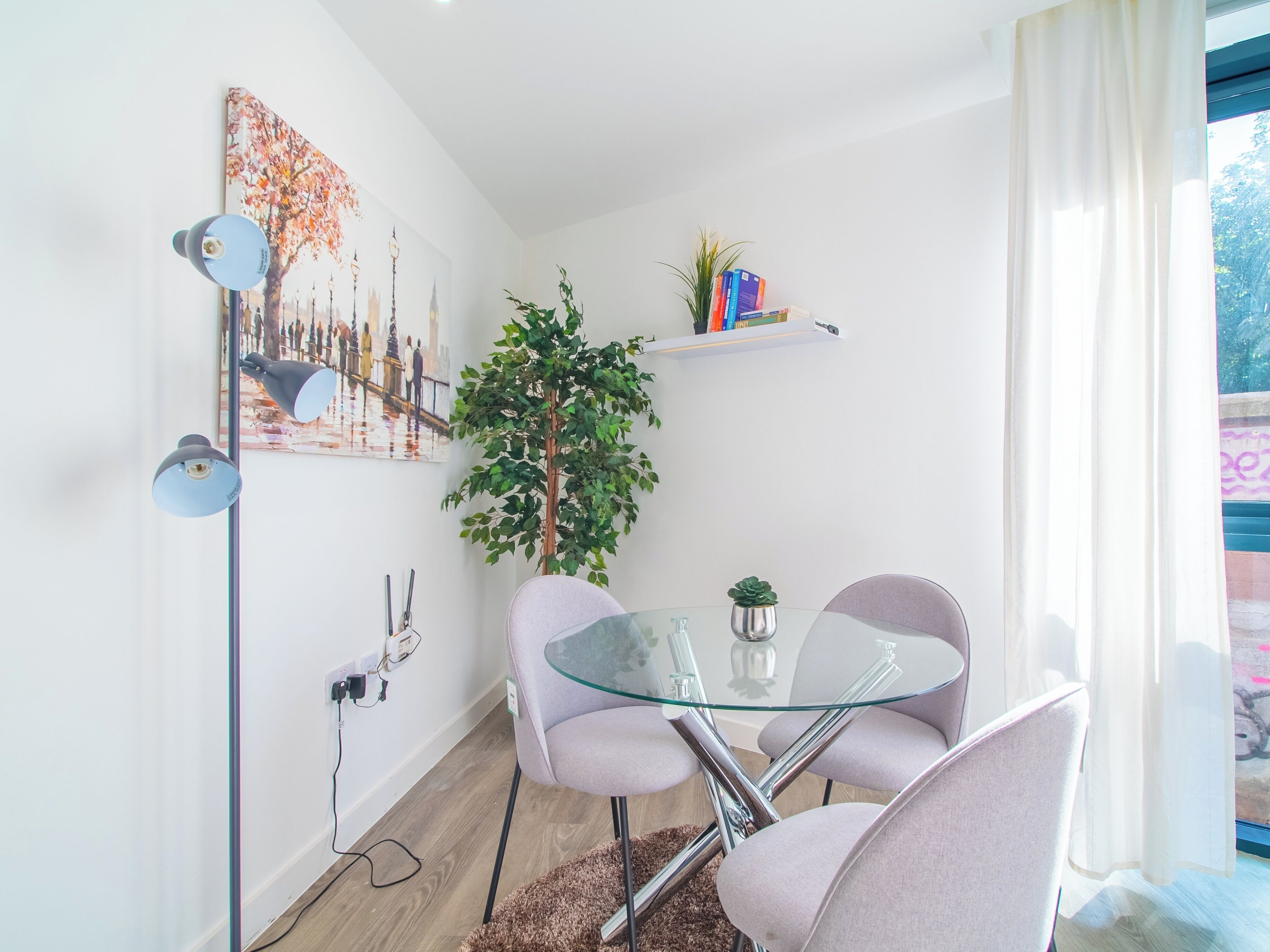 Un comedor moderno con una mesa de vidrio, dos sillas, una planta en maceta y un cuadro de un paisaje urbano en la pared.