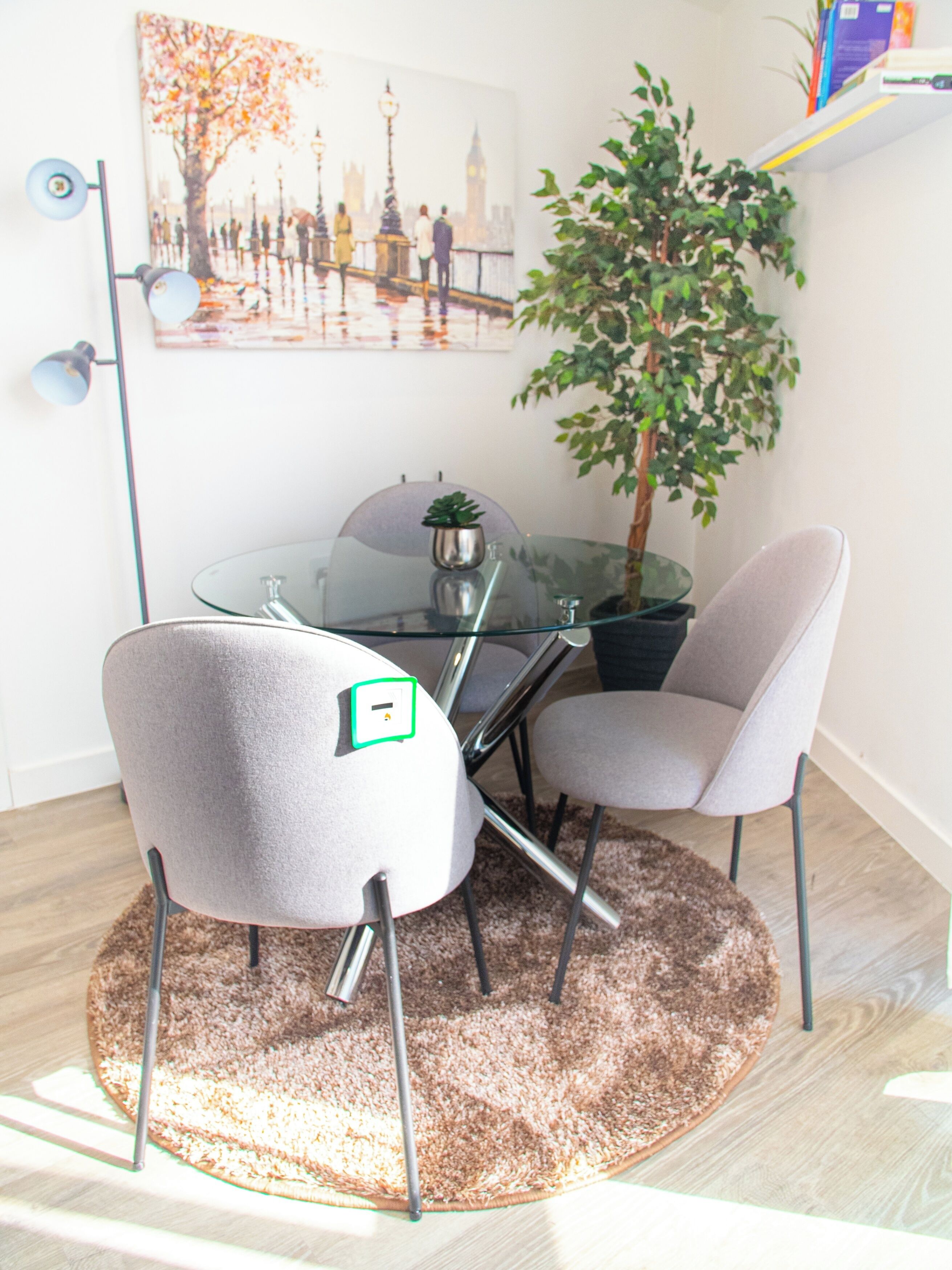 Un espace repas moderne avec une table en verre, deux chaises grises, une plante en pot et un tableau encadré représentant un paysage urbain accroché au mur.