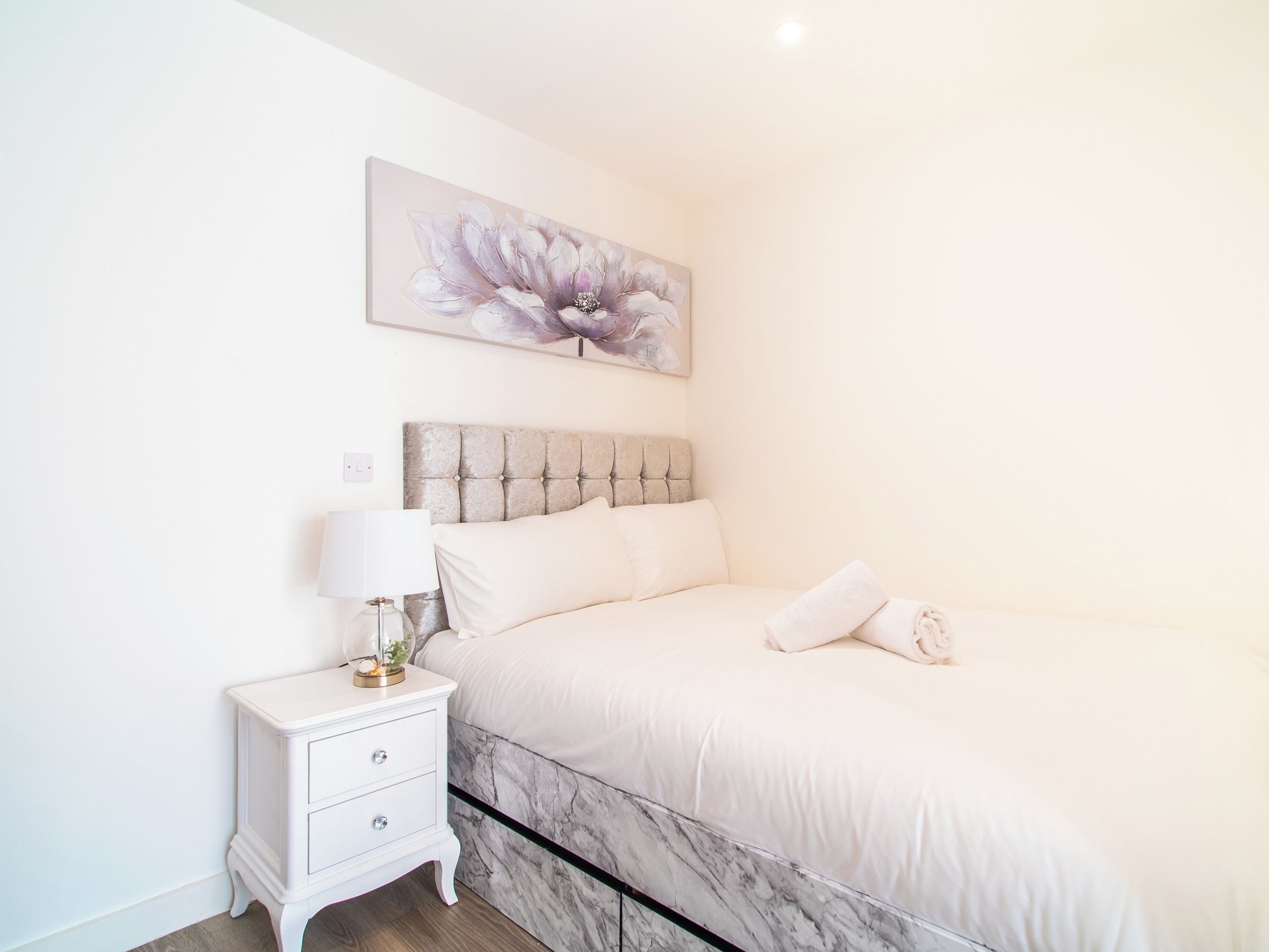 A bedroom with a marble headboard, a white bed, a bedside table with a lamp, and a floral painting on the wall.