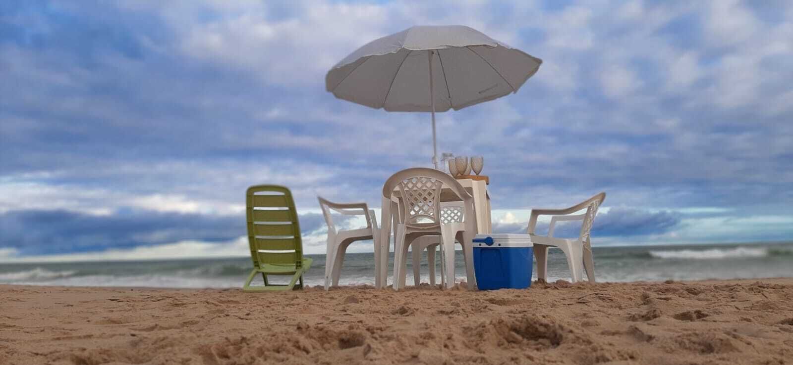 A beach setting with a white umbrella, plastic chairs, and a blue cooler on the sand.
