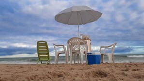 Une plage avec un parasol blanc, des chaises en plastique et une glacière bleue posée sur le sable.