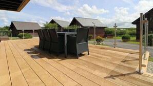 A wooden deck with a table and chairs, overlooking a row of houses with sloped roofs. - Four bedroom holiday lodge with onsite watersports. (Saint Columb Major)