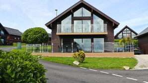 A two-story wooden house with a balcony, a well-maintained lawn, and a clear sky. - Four bedroom holiday lodge with onsite watersports. (Saint Columb Major)