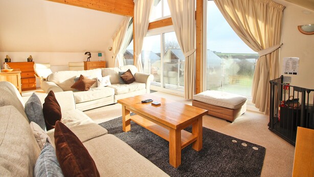 A spacious living room with a wooden ceiling, a large window with curtains, a sofa set, a coffee table, and a view of the outside. - Four bedroom holiday lodge with onsite watersports. (Saint Columb Major)