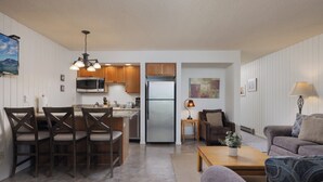 Una cocina moderna con armarios de madera, un microondas y un refrigerador. Hay un comedor con una mesa y sillas, y una sala de estar con un sofá y una mesita de centro.