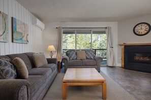 Un salon avec un canapé, un fauteuil et une table basse. On y trouve une cheminée et une horloge au mur.