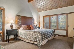 A bedroom with a large bed, two bedside tables, a nightstand, a wooden headboard, a window with blinds, and a stone accent wall.