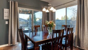 A dining area with a wooden table and chairs, a vase with flowers, and a view of a backyard through large windows. - 4BR Bend Home Near Mt Bachelor and River Trails (Bend)