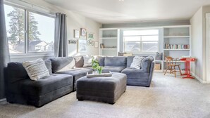 A living room with a sectional sofa, ottoman, and a small table with a tray. There is a bookshelf and a window with curtains. - 4BR Bend Home Near Mt Bachelor and River Trails (Bend)