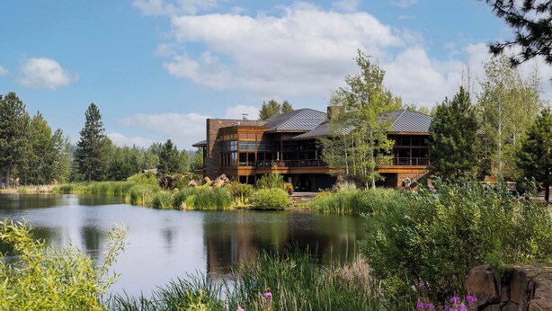 Una casa grande y moderna frente a un lago, rodeada de abundante vegetación y árboles.