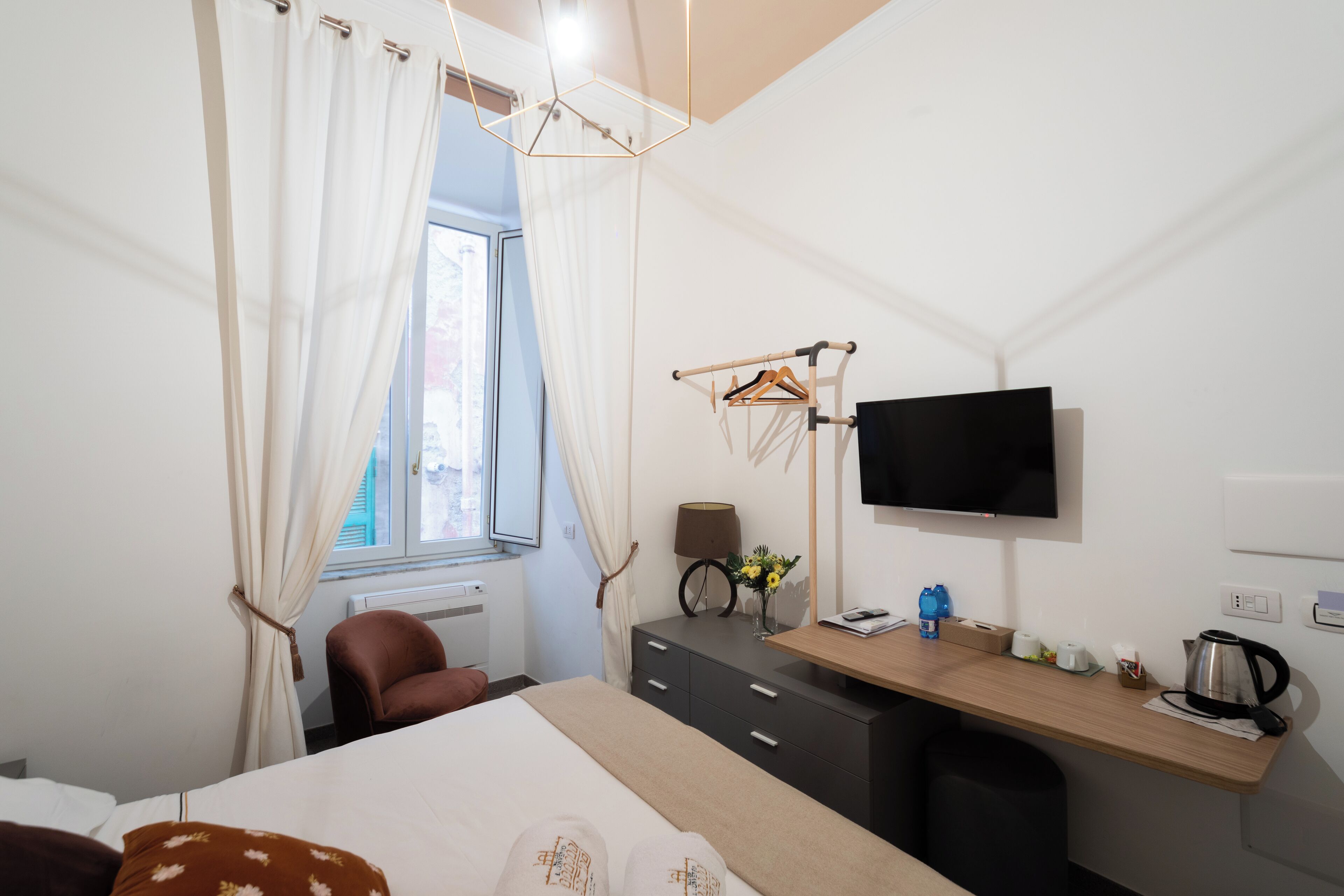 A modern hotel room with a bed, a desk with a flat-screen TV, a chair, a dresser, and a window with curtains.