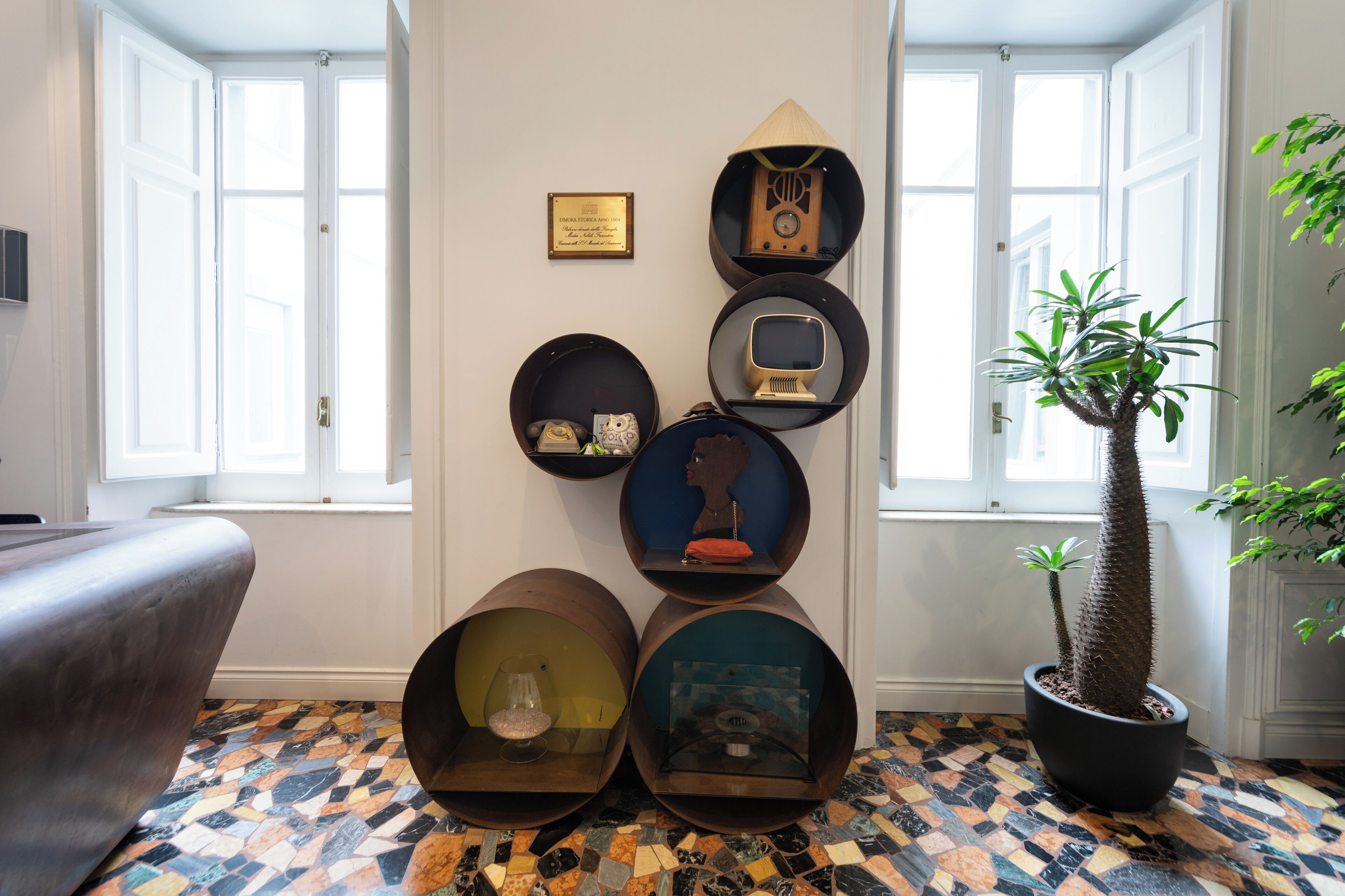 A room with a patterned floor, a potted plant, and wall-mounted shelves with various items.