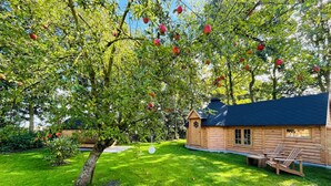A wooden cabin with a sloped roof, surrounded by a well-maintained lawn and a tree bearing red fruits. - B&B Voor De Wind (Slootdorp)