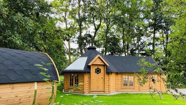 A wooden cabin with a pitched roof and a large window, surrounded by a well-maintained lawn and trees. - B&B Voor De Wind (Slootdorp)