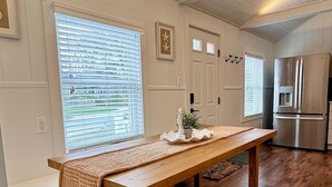 A kitchen with a wooden table, a refrigerator, and a window with blinds. - Charming Cape Cod Getaway- EV Charger, Mini Golf, Walk to The Beach, Pets Okay! (Yarmouth)