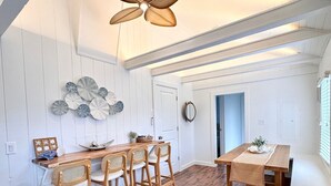 A dining area with a wooden table and chairs, a ceiling fan, and a decorative wall piece. - Charming Cape Cod Getaway- EV Charger, Mini Golf, Walk to The Beach, Pets Okay! (Yarmouth)