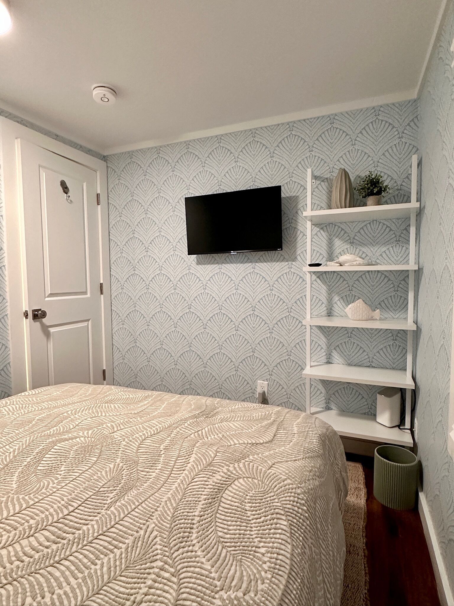 A bedroom with a bed, a TV mounted on the wall, a white door, and a shelving unit with decorative items.
