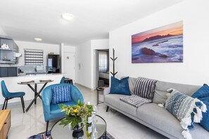 Un salon moderne avec un canapé, des fauteuils, une table basse et un tableau représentant un coucher de soleil sur l’océan.