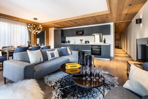A modern living room with a grey sofa, a coffee table with wine glasses, and a dining area in the background. - Wifi, 1st floor, view mountain, sauna, hammam, terrace, fireplace or stove, tv, 91m², Méribel (Méribel - Les Allues)