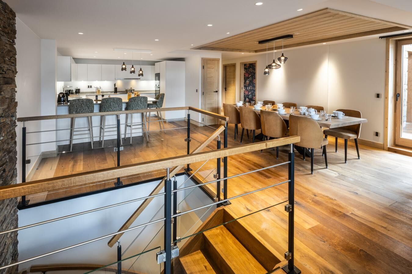 A modern kitchen with white cabinets, a dining area with a wooden table and chairs, and a staircase with glass railings.