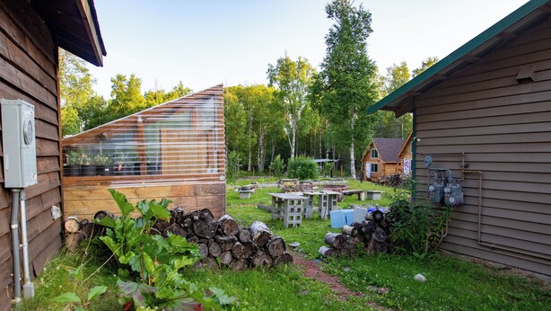 A wooden cabin with a glass greenhouse, a wooden deck, and a garden area with logs and plants. - Quaint Rural Cabin Near Hatcher Pass (Wasilla)