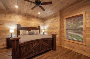 Ein Schlafzimmer einer Holzhütte mit einem Bett, zwei Nachttischlampen, einem Deckenventilator und einem Fenster mit Jalousien.