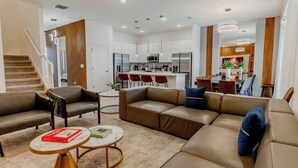 A modern living room with a sofa, armchairs, and a coffee table. There is a kitchen in the background with a dining area.
