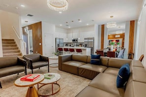 Una sala de estar moderna con un sofá, sillones y una mesa de centro. Se observa una cocina al fondo con un comedor.