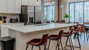 A modern kitchen with white cabinets, stainless steel appliances, a central island with bar stools, and large windows offering natural light.