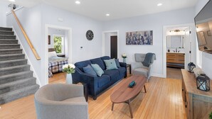 Una sala de estar moderna con una escalera, un sofá azul, una mesa de centro de madera y una pintura de un paisaje urbano.