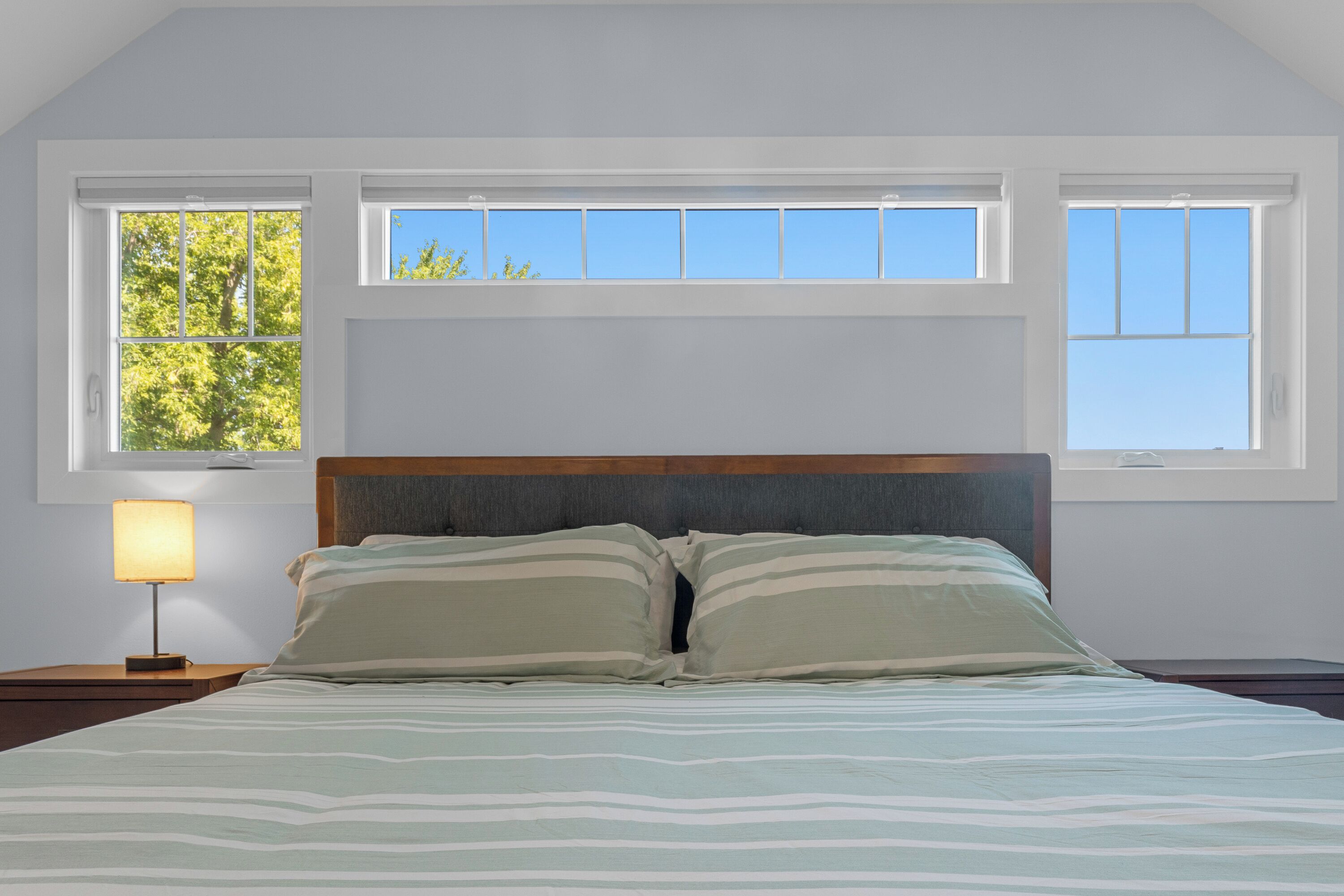 Un dormitorio con cama, dos ventanas, una mesita de noche con lámpara y vistas a árboles.