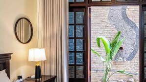 Habitación de hotel con cama, mesita de noche con lámpara, espejo, ventana con vista a un muro de piedra y una planta verde.