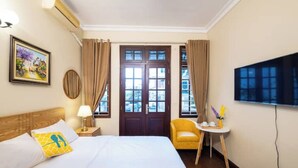 Habitación de hotel con cama, una silla, una mesita con un florero, un cuadro en la pared y un televisor montado en la pared.