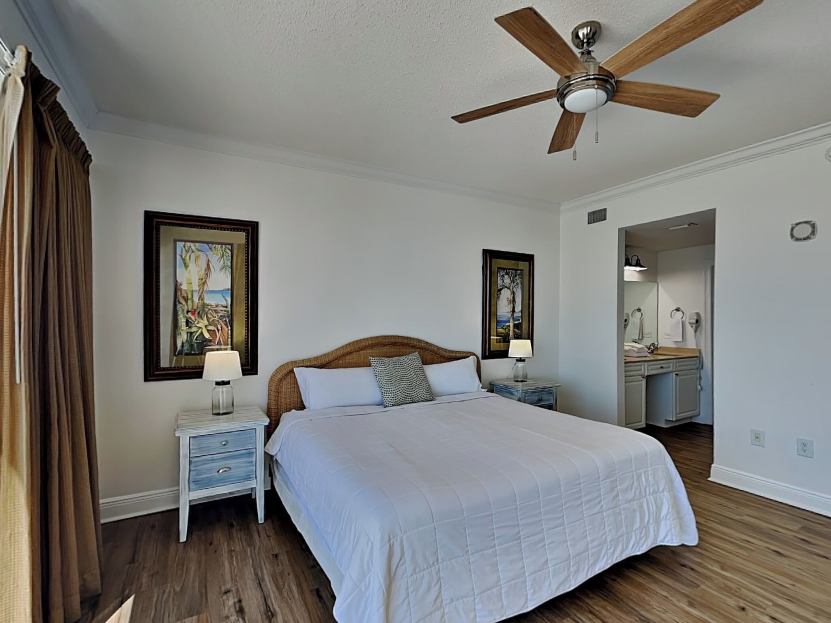 A bedroom with a large bed, a ceiling fan, two bedside tables with lamps, and a bathroom visible through an open door.