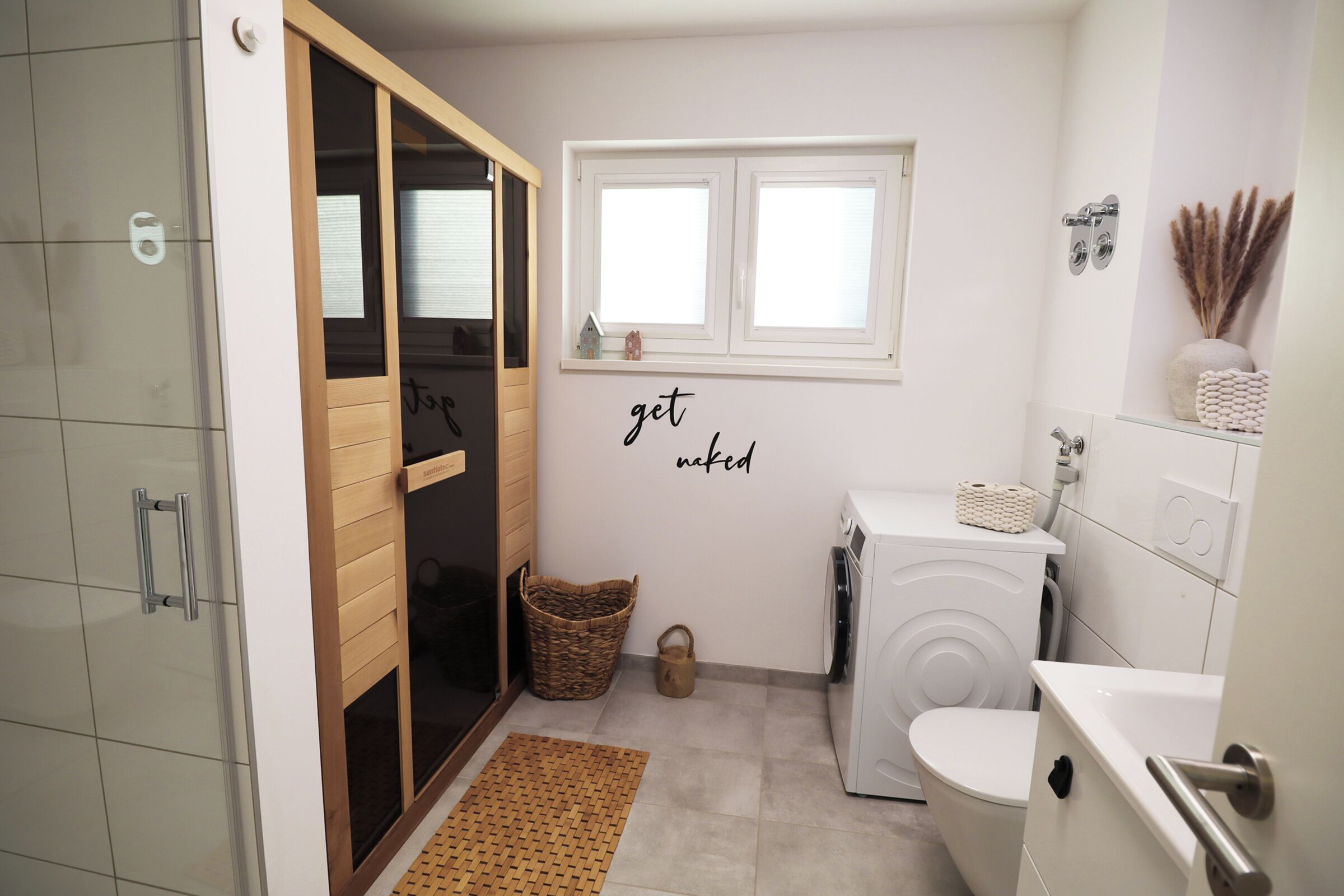 A compact bathroom with a washing machine, a toilet, a small window, and a wall-mounted shelf with decorative items.