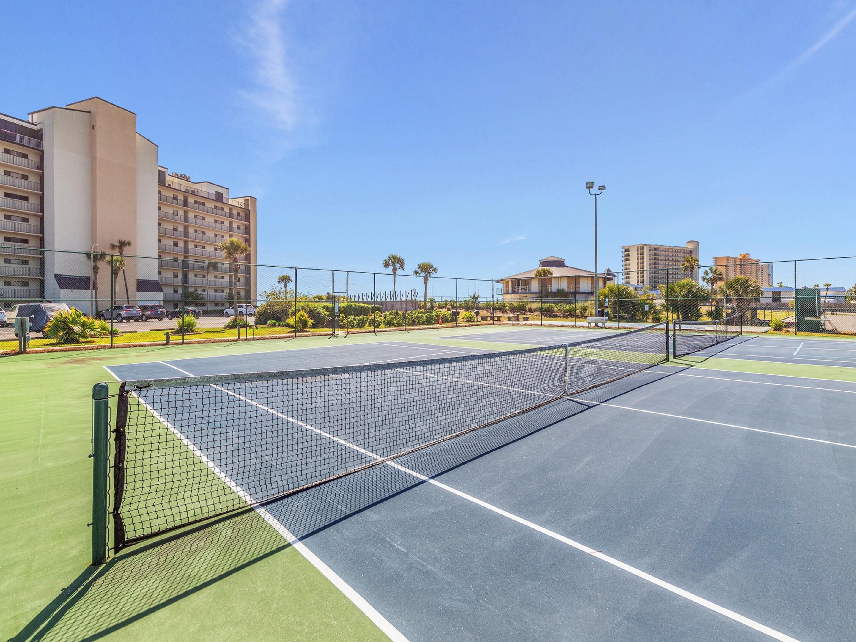 Ein Tennisplatz mit Netz, umzäunt und neben einem Gebäude mit mehreren Balkonen.