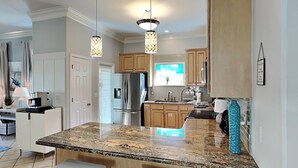 A modern kitchen with granite countertops, stainless steel appliances, and wooden cabinets. There are two white bar stools and pendant lights above the island. - Paradise Escape by Southern Vacation Rentals (Destin)