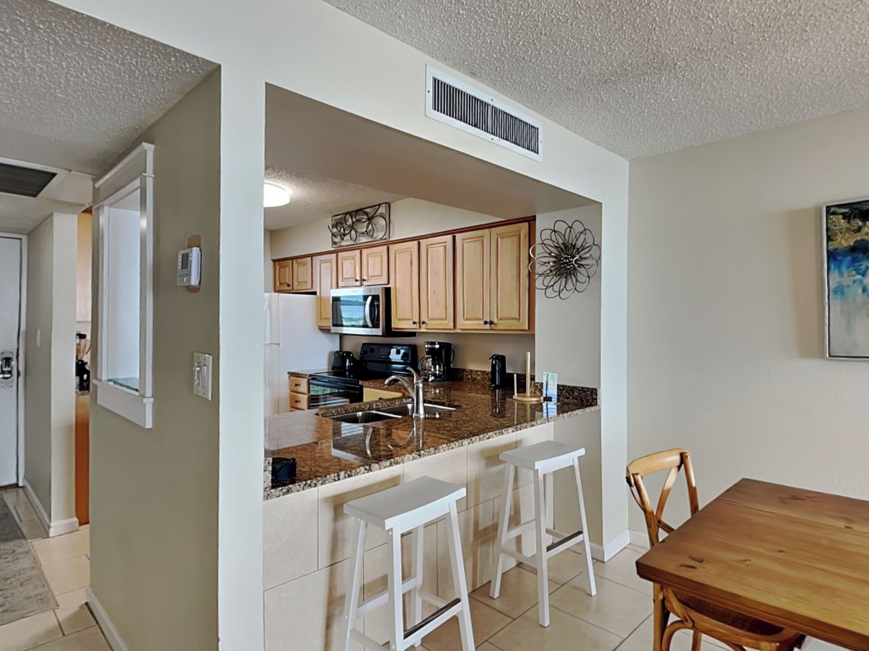 A modern kitchen with a central island, wooden cabinets, and a microwave. There are two white bar stools and a wooden dining table with chairs.