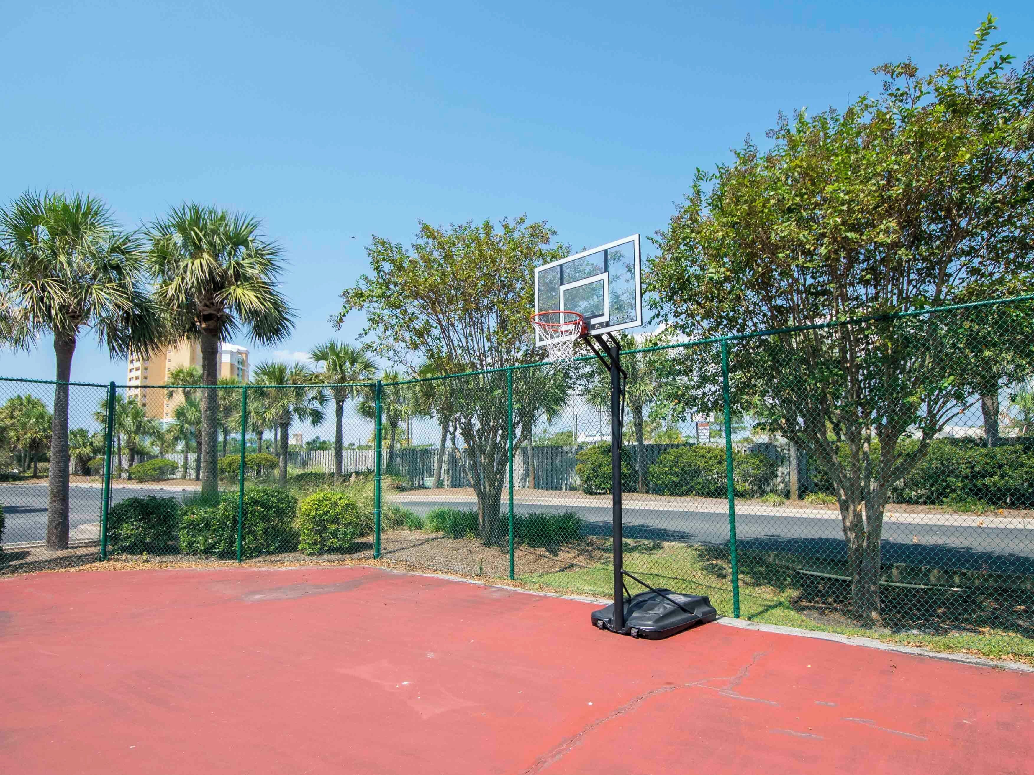 Ein Basketballplatz im Freien mit Korb, umgeben von einem Maschendrahtzaun, Palmen und blauem Himmel.