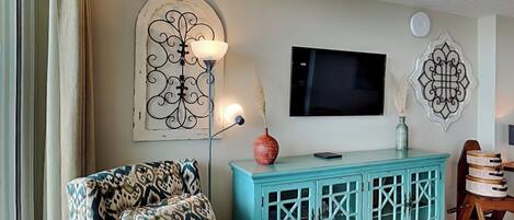 Un soggiorno con una poltrona a fantasia, un mobiletti color petrolio, una televisione a schermo piatto, decorazioni a parete e un tavolino con un vaso.