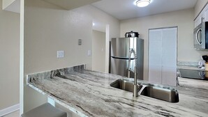 A kitchen with a marble countertop, stainless steel sink, and a refrigerator.
