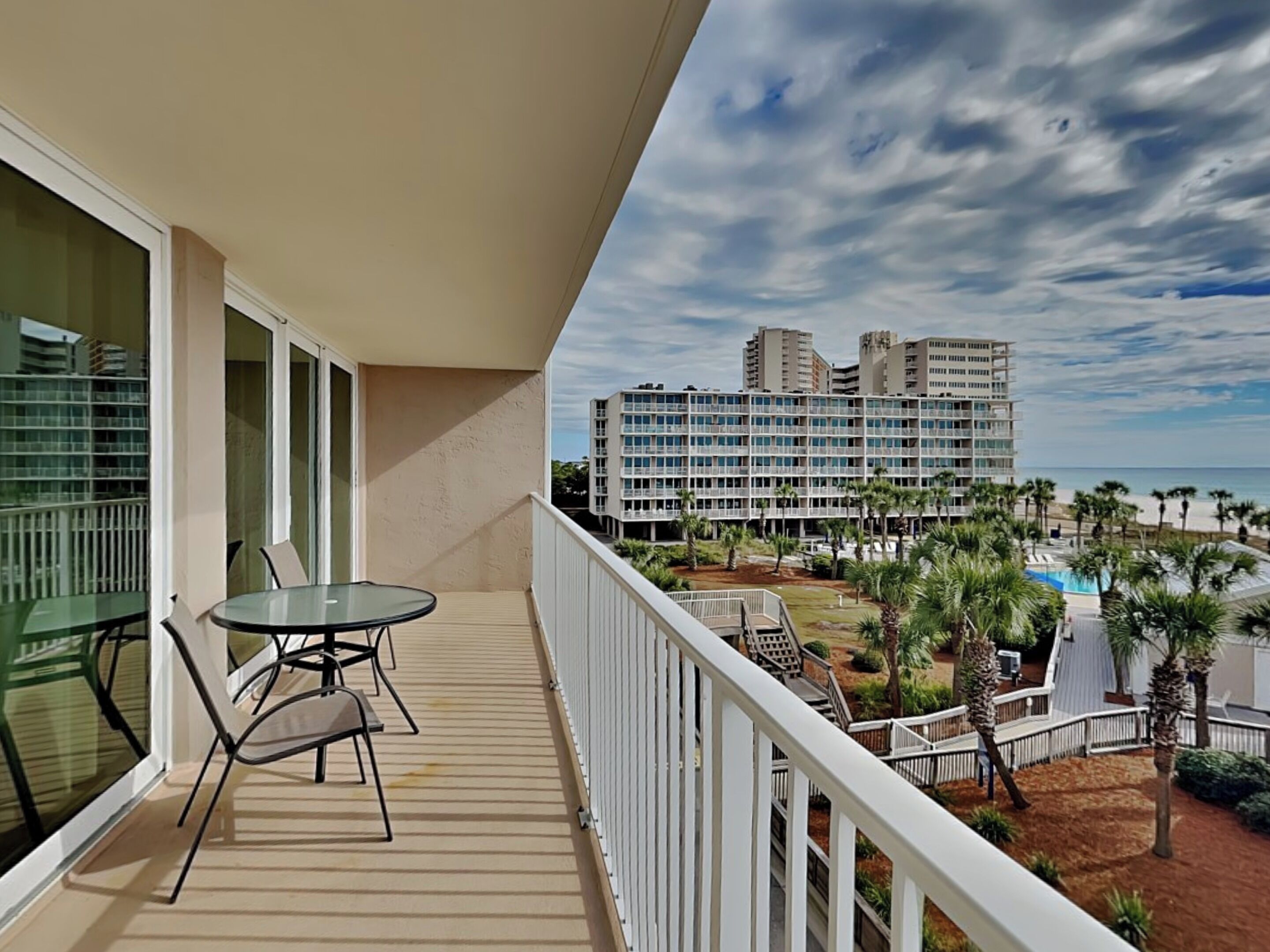 Un balcon avec une table et des chaises en verre, donnant sur un espace piscine agrémenté de palmiers et sur un hôtel en bord de mer en arrière-plan.