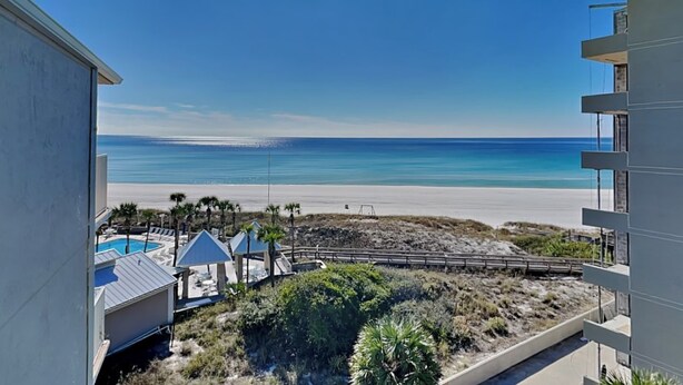 A beachfront view from a balcony overlooking a pool area with lounge chairs and umbrellas, a sandy beach, and a clear sky.